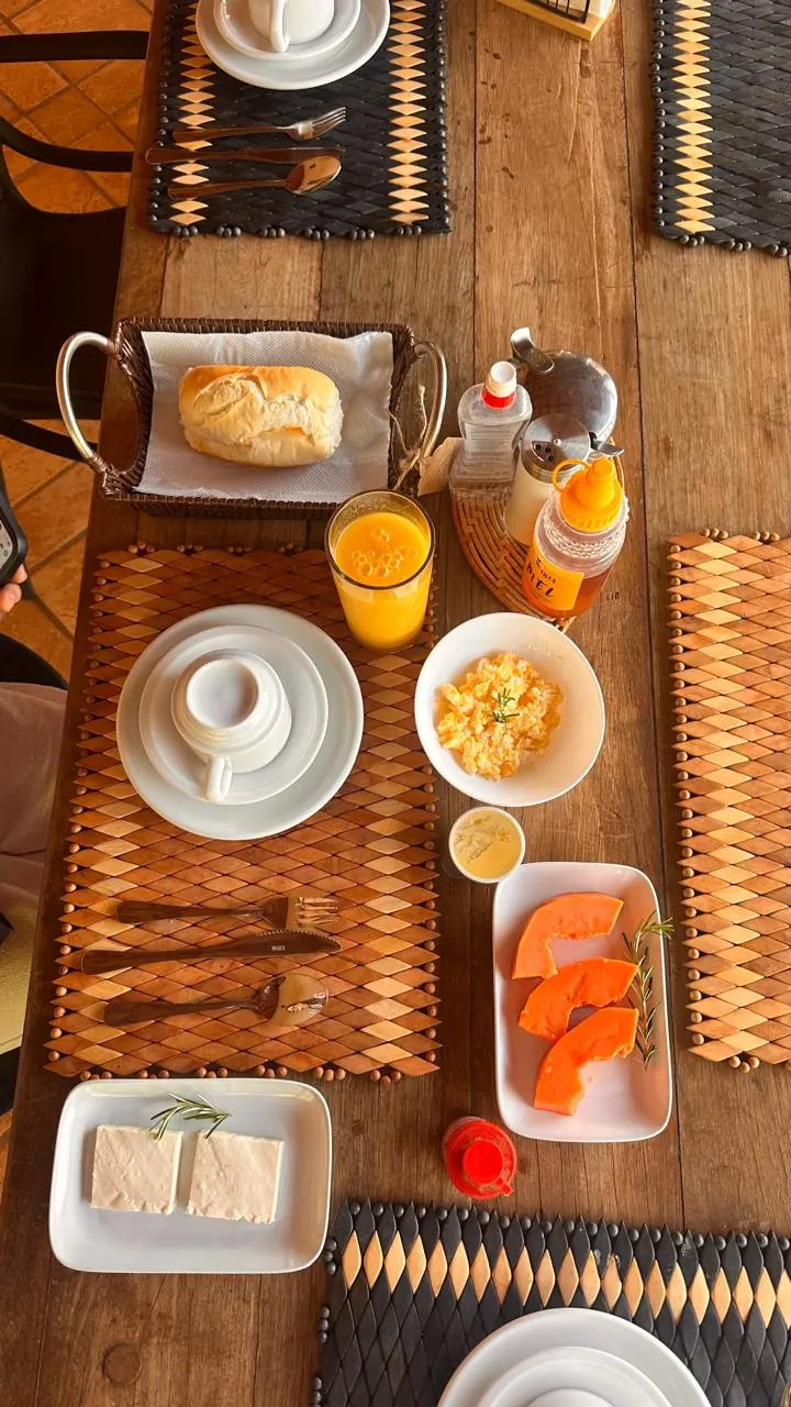 Mesa de madeira posta, vista de cima, com xícara, pão, suco, ovos mexidos, mamão e queijo
