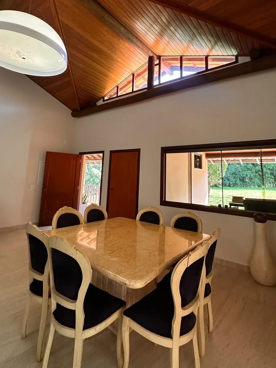 Mesa de jantar de mármore com oito cadeiras, em sala de pé-direito alto com forro de madeira e janela para o jardim